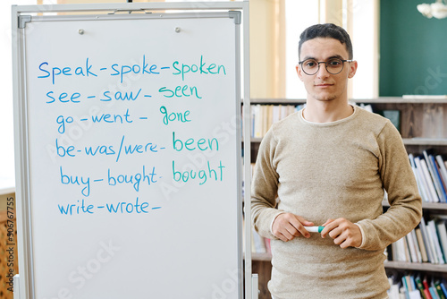 Horizontal medium portrait of young Middle-Eastern student standing at whiteboard with irregular verbs written on it looking at camera photo