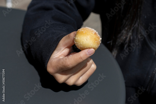 Kid's hand holding a cinnamon sugar dusted brioche donut hole on an outdoor table in the afternoon sun.