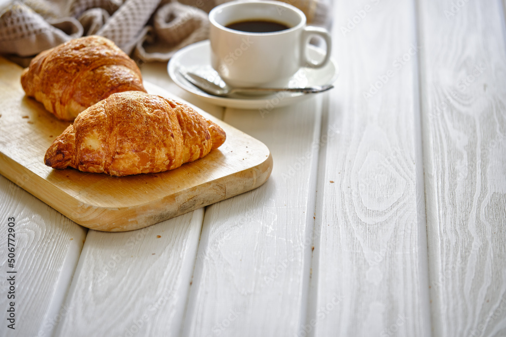A crispy croissant with almond filling and hot black coffee on a wooden board on a light concrete table.