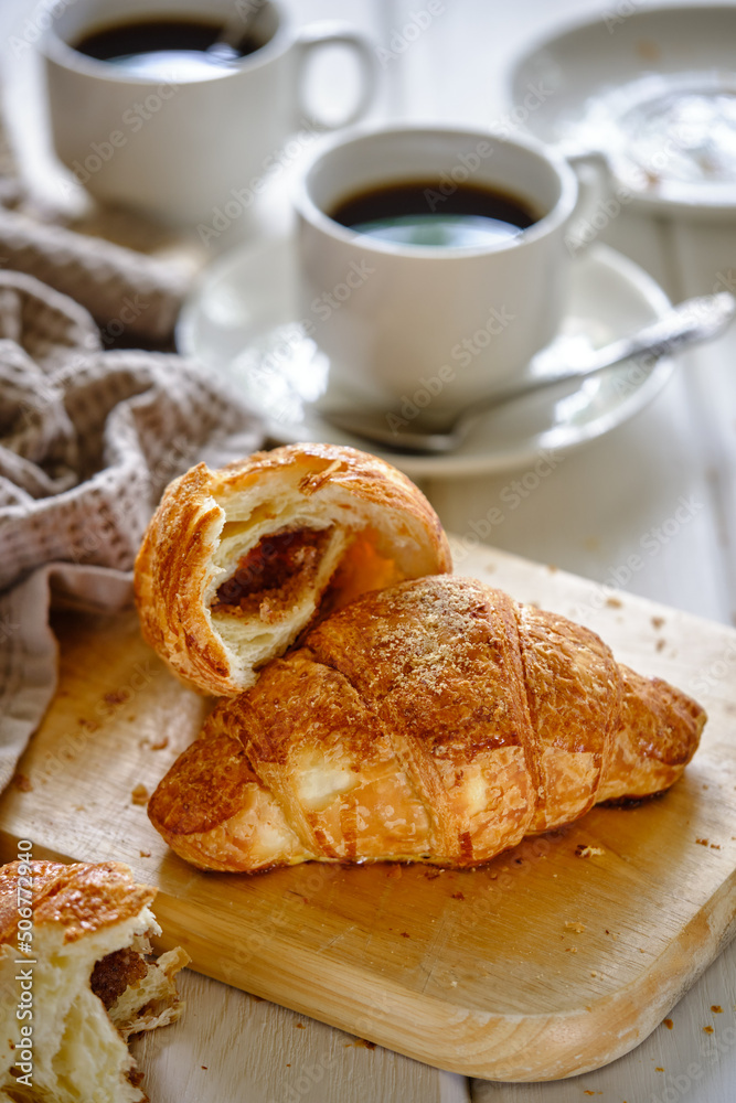 A crispy croissant with almond filling and hot black coffee on a wooden board on a light concrete table.