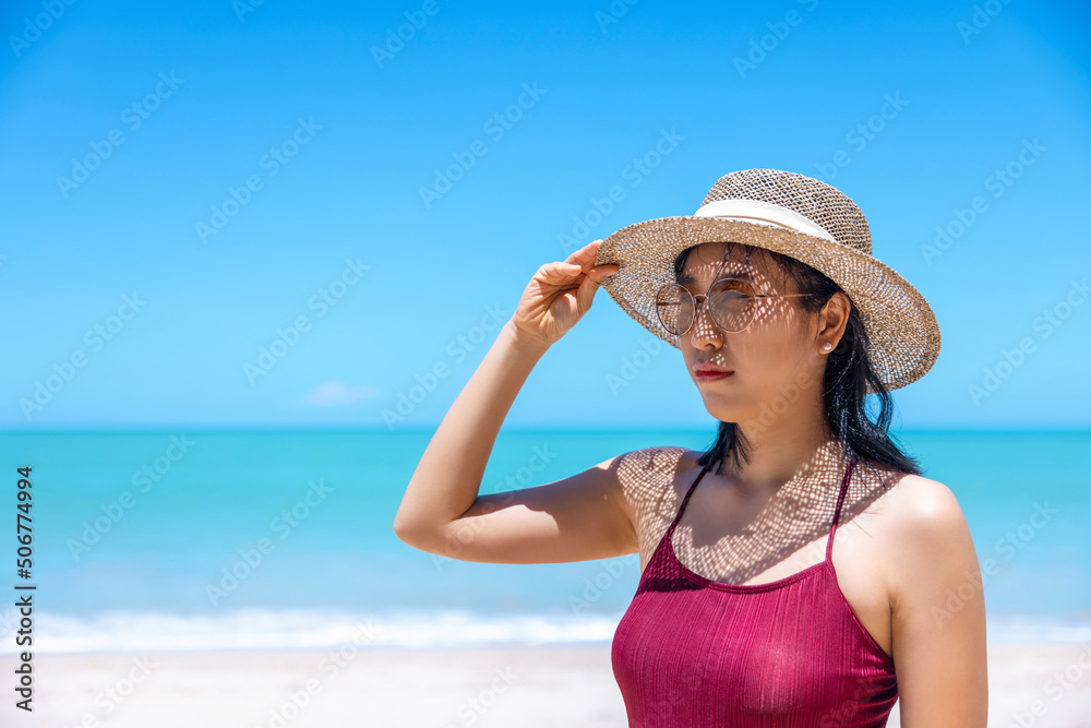 Young women in bikini and straw hat stand on tropical beach enjoying looking view of beach ocean on hot summer day. Blue sea in background. Khao Lak, Phang Nga, Thailand. Summer vacation concept