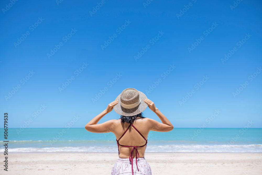Young women in bikini and straw hat stand on tropical beach enjoying looking view of beach ocean on hot summer day. Blue sea in background. Khao Lak, Phang Nga, Thailand. Summer vacation concept Young women in bikini and straw hat stand on tropical beach enjoying looking view of beach ocean on hot summer day. Blue sea in background. Khao Lak, Phang Nga, Thailand. Summer vacation concept