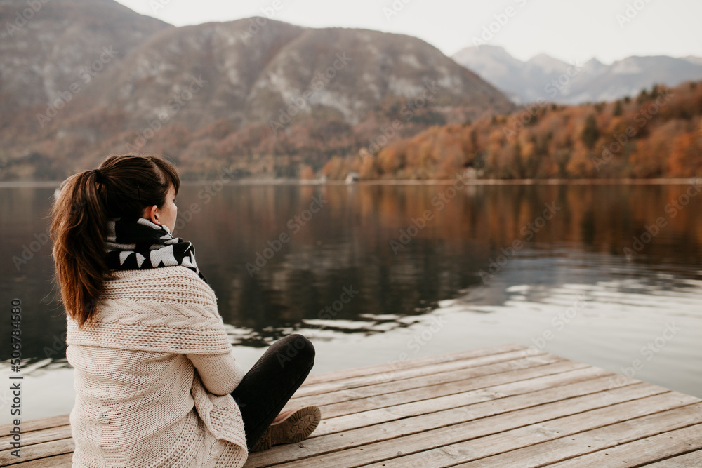 A woman is sitting on a wooden pier and meditating. She is sitting by lake Bohinj in Slovenia. Stunning scenery, beautiful autumn landscape. Perfect place to relax. Lakeside and mountains. Copy space.