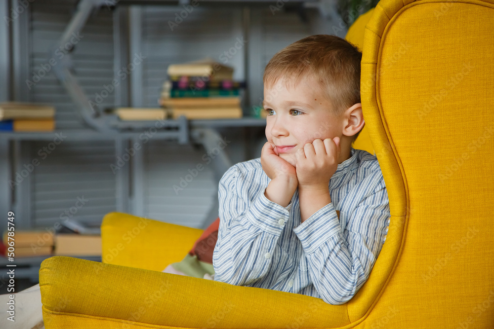 A little blond boy in a yellow armchair looks away with a smile. Space for text. International Children's Day A little blond boy in a yellow armchair looks away with a smile. Space for text. International Children's Day
