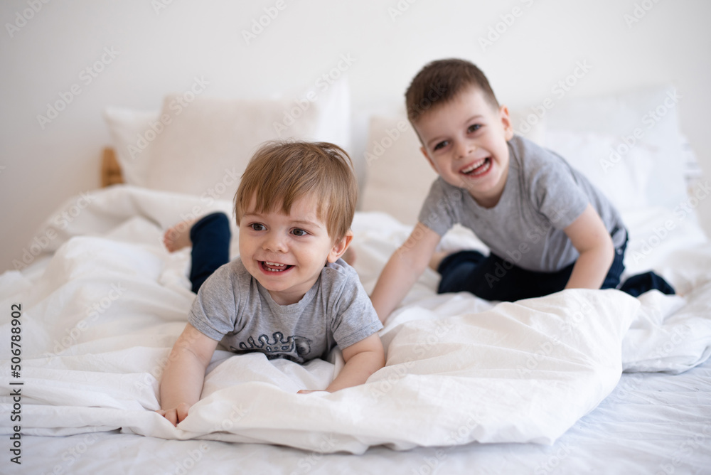 Smiling two boys in gray t-shirts lie and playing on white blanket on bed. Cheerful and happy childhood in family. Exercise and play good for health. Together joyful and interesting. Smiling two boys in gray t-shirts lie and playing on white blanket on bed. Cheerful and happy childhood in family. Exercise and play good for health. Together joyful and interesting.