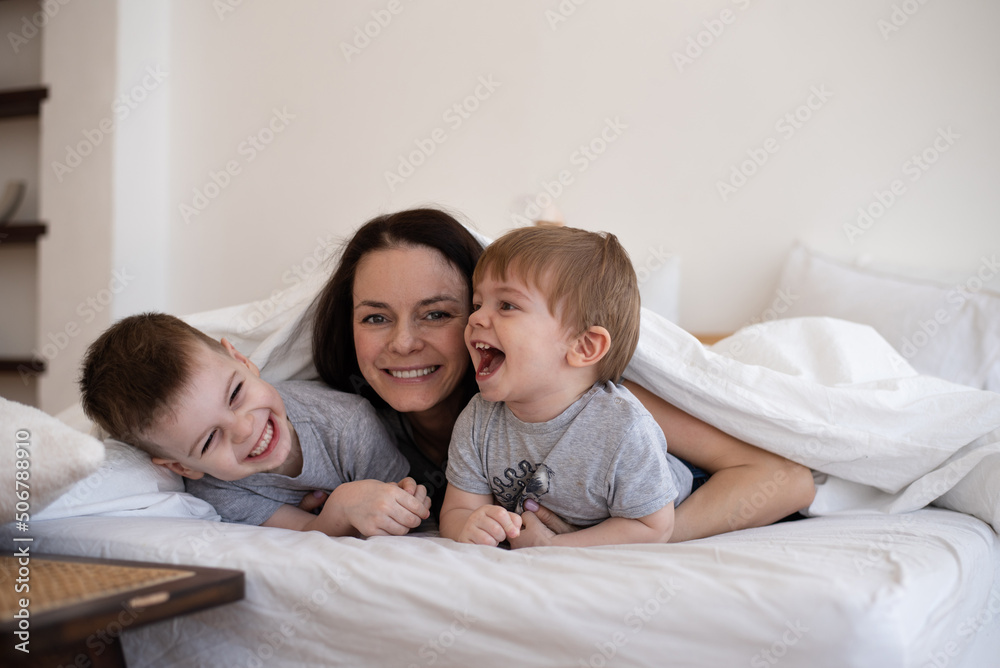 Happy family: mom and two sons lying in bed under blanket at home. Woman with children have fun in bedroom. Love, smiles and good mood brings together family.