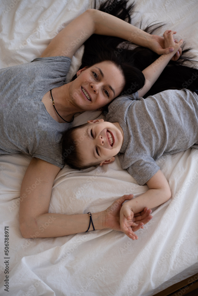 Mom and son lying and smiling in bed on white blanket at home. Woman with child have fun in bedroom. enjoy and happiness together.