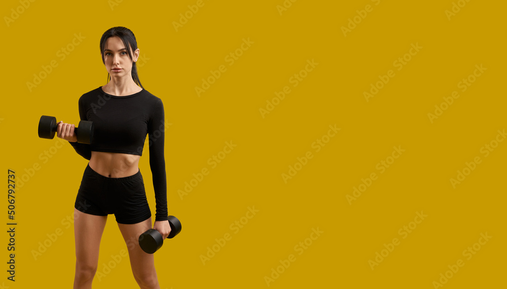 Front view of pretty slim girl in sportswear working out, training, exercising. Brunette female with ponytail standing, raising dumbbells, looking at camera. Concept of youth. Front view of pretty slim girl in sportswear working out, training, exercising. Brunette female with ponytail standing, raising dumbbells, looking at camera. Concept of youth.
