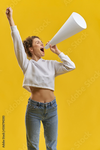 Side view of young female standing, speaking in megaphone. Pretty girl raising hand, shouting, crying, encouraging, wearing khudi, jeans. Isolated on yellow studio background.