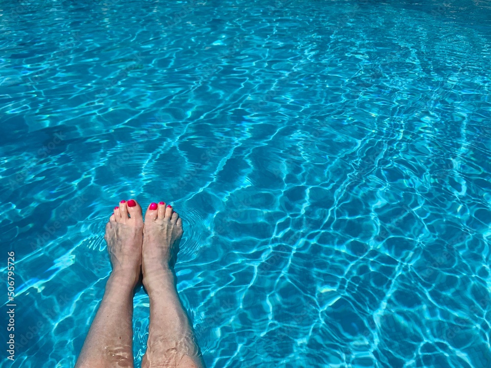 Middle aged woman's legs, with red nail polish on toenails, splashing feet in turquoise blue waters of swimming pool. copy space