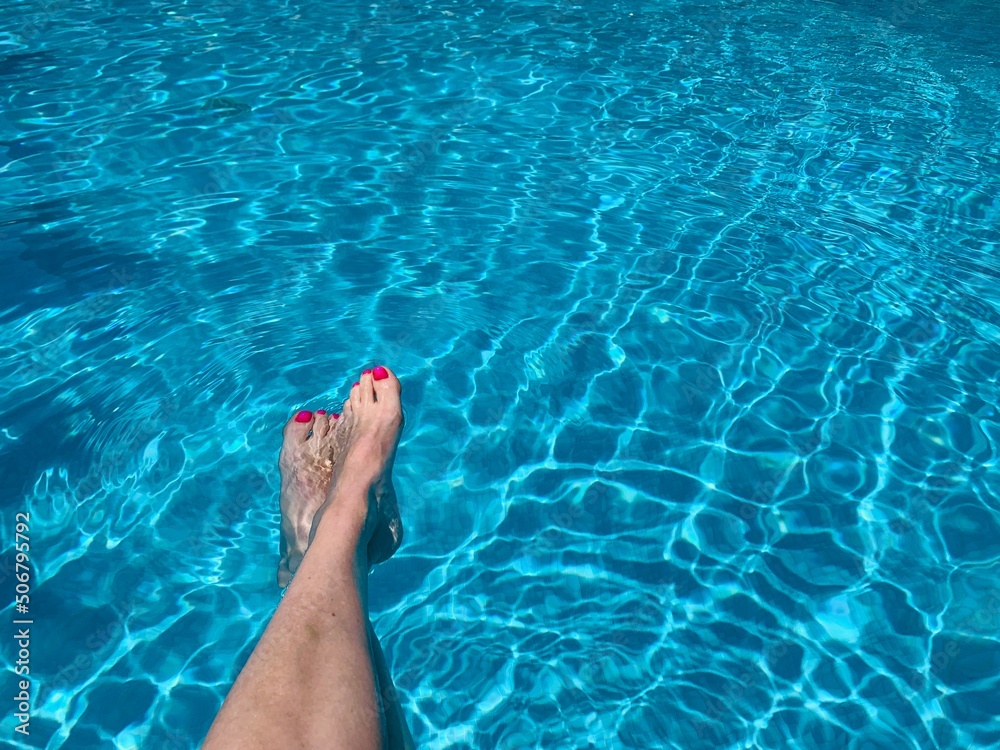 Middle aged woman's legs, with red nail polish on toenails, splashing feet in turquoise blue waters of swimming pool. copy space
