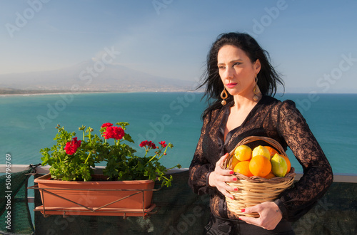 View of a typical mediterranean woman holding a wicker basket full of oranges and lemons in a panoramic balcony in Sicily, with blue sea and Mount Etna in the background