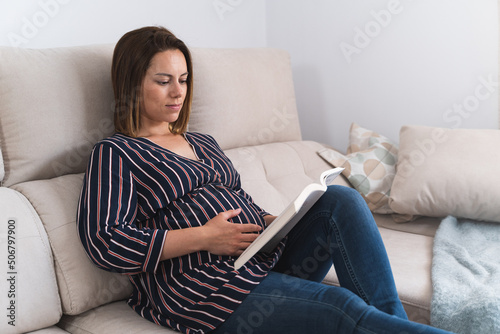 Profile of a pregnant woman touching the belly while reading a book on the sofa at home