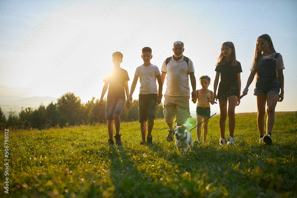 Front view of children with old man and dog walking in mountains. Male with gray hair and beard with kids leding jack russell terrier, going forward at sunset. Concept of friendship. Front view of children with old man and dog walking in mountains. Male with gray hair and beard with kids leding jack russell terrier, going forward at sunset. Concept of friendship.
