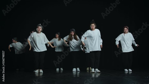 Teenagers dance a rehearsed dance. They make synchronous, precise movements. People are dressed the same. They have white tops and black bottoms. There are both boys and girls here