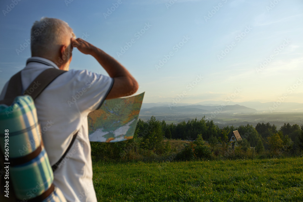 Back panoramic view of old man with gray hair and rucksack holding map in mountains. Male hiding from sun, holding by hand, looking at landscape in hills. Concept of traveling.