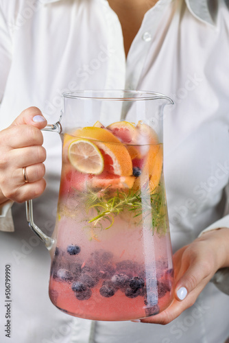 A jug with a refreshing organic soft drink with lime, blueberry, grapefruit and rosemary in female hands close-up photo