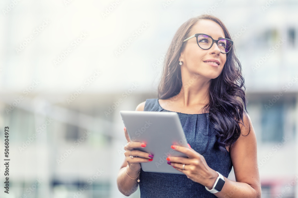Businesswoman in glasses looks aside standing outdoors holding a tablet Businesswoman in glasses looks aside standing outdoors holding a tablet