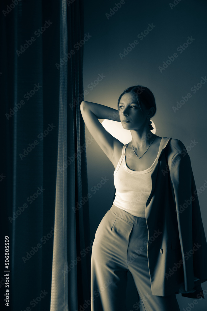 Portrait of young woman in beige suit standing in room next to the windows curtain Portrait of young woman in beige suit standing in room next to the windows curtain