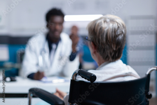 Selective focus on sick senior patient with disability talking with physician about illness examination appointment. Woman in wheelchair conversating with doctor about health check and prescribed