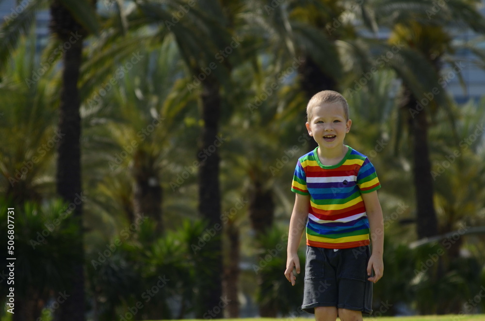 A little boy in colorful rainbow t-shirt. In the background is a palm avenue. A child rejoices in the summer Israel May 2022space for text. A little boy in colorful rainbow t-shirt. In the background is a palm avenue. A child rejoices in the summer Israel May 2022space for text.