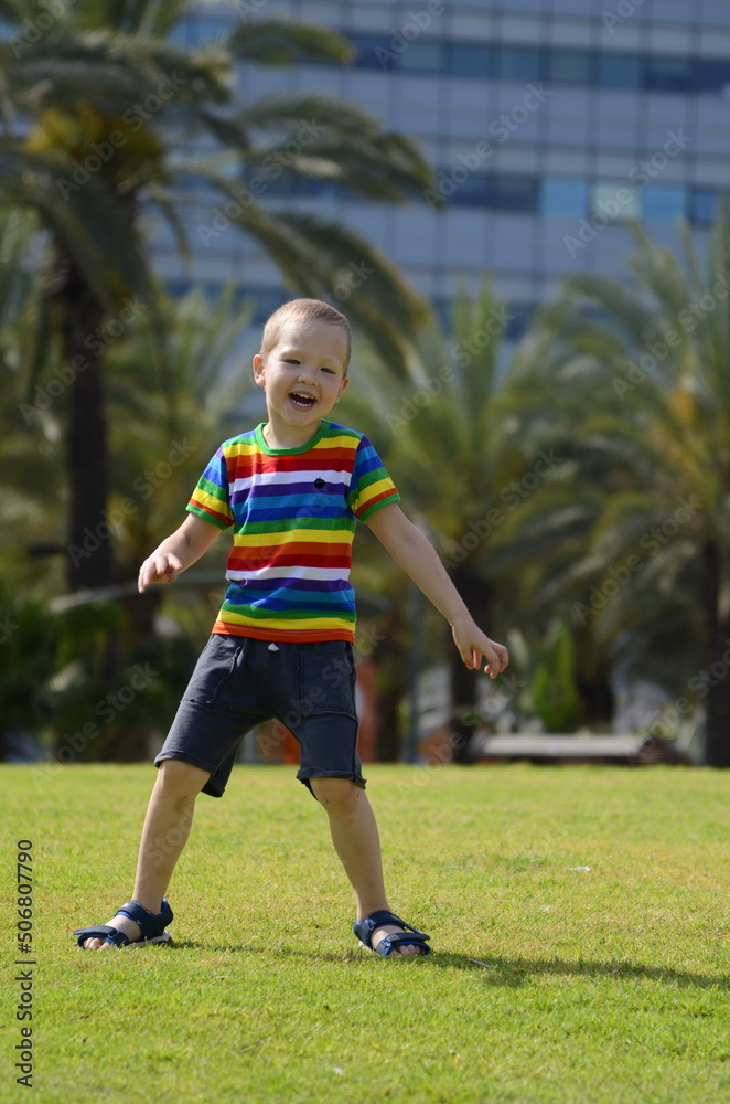 A little boy jumps on a green meadow in the park. In the background is a palm avenue. A child in a multi-colored T-shirt rejoices in the summer
