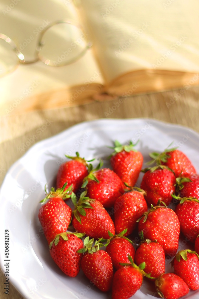 Lilac plate full of fresh strawberries, open book and reading glasses on wooden table. Selective focus. Lilac plate full of fresh strawberries, open book and reading glasses on wooden table. Selective focus.