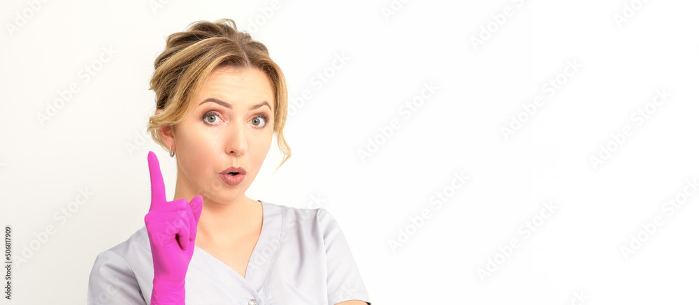 Young caucasian woman doctor wearing pink gloves keeping index finger pointed upwards making a gesture with index finger. I have an idea Young caucasian woman doctor wearing pink gloves keeping index finger pointed upwards making a gesture with index finger. I have an idea