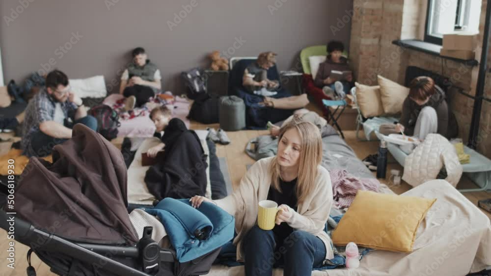 Medium long high angle of young Caucasian mother rocking unrecognizable baby in stroller, drinking hot coffee, sitting on cot in foreground of blurred refugees in shelter at daytime