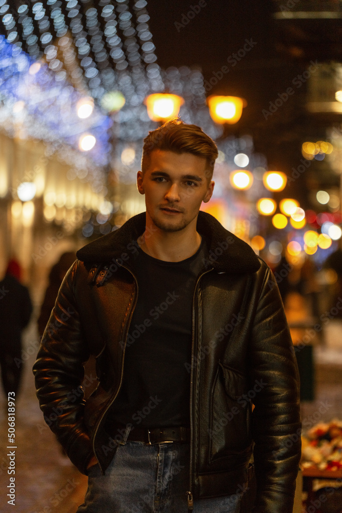 Fashionable handsome man with hair in a fashionable black clothes with leather jacket and jeans walking on a night street in the city Fashionable handsome man with hair in a fashionable black clothes with leather jacket and jeans walking on a night street in the city