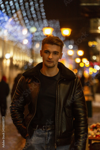 Fashionable handsome man with hair in a fashionable black clothes with leather jacket and jeans walking on a night street in the city