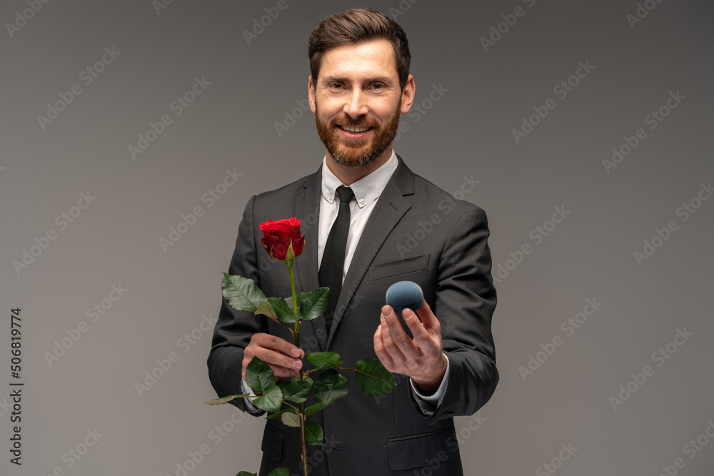Satisfied happy man dressed in formal suit holding box with an engagement ring and stretching it to the camera isolated over grey background Satisfied happy man dressed in formal suit holding box with an engagement ring and stretching it to the camera isolated over grey background