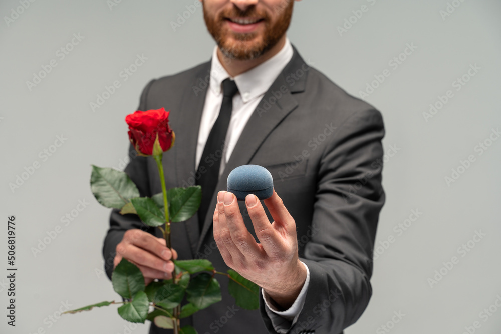 Portrait of a satisfied happy man dressed in formal suit holding box with an engagement ring and stretching it to the camera isolated over grey background Portrait of a satisfied happy man dressed in formal suit holding box with an engagement ring and stretching it to the camera isolated over grey background