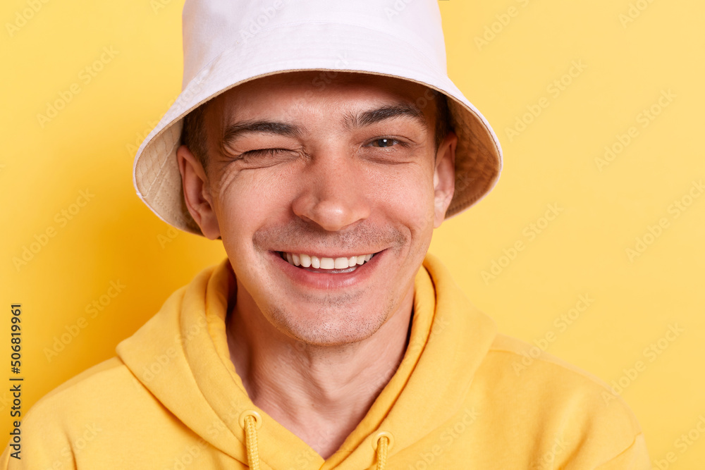 Indoor shot of childish man wearing casual style hoodie and panama posing isolated over yellow background, standing and winking playfully, positive expression, flirting. Indoor shot of childish man wearing casual style hoodie and panama posing isolated over yellow background, standing and winking playfully, positive expression, flirting.