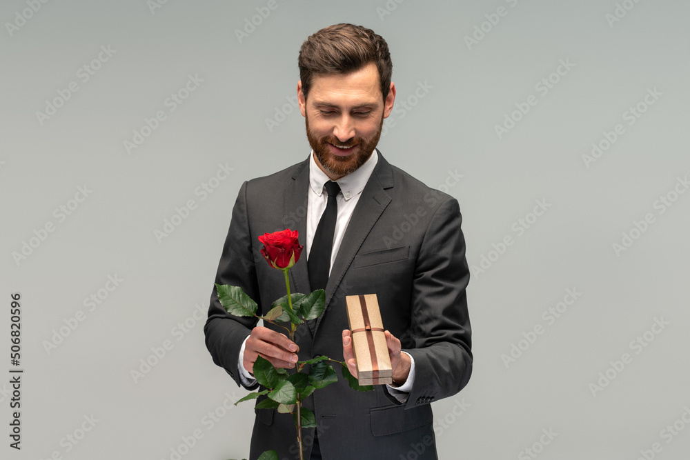 Handsome bearded brunette man in formal suit standing, holding red rose flower and looking at box with present. Indoor studio shot isolated on grey background Handsome bearded brunette man in formal suit standing, holding red rose flower and looking at box with present. Indoor studio shot isolated on grey background