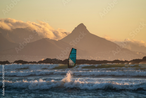 Windsurfer in focus surfing waves during the beautiful golden hour in Cape Town with Table Mountain in the back. Big Bay beach is an amazing place for sunset surf and windsurfing sessions. 