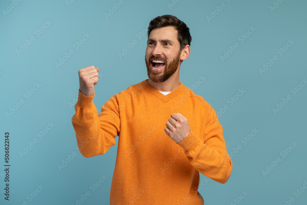 Extremely happy man in orange outfit raised arm gesturing yes i did it, I'm winner, screaming loud for joy feeling energetic and lively. Isolated on blue background Extremely happy man in orange outfit raised arm gesturing yes i did it, I'm winner, screaming loud for joy feeling energetic and lively. Isolated on blue background