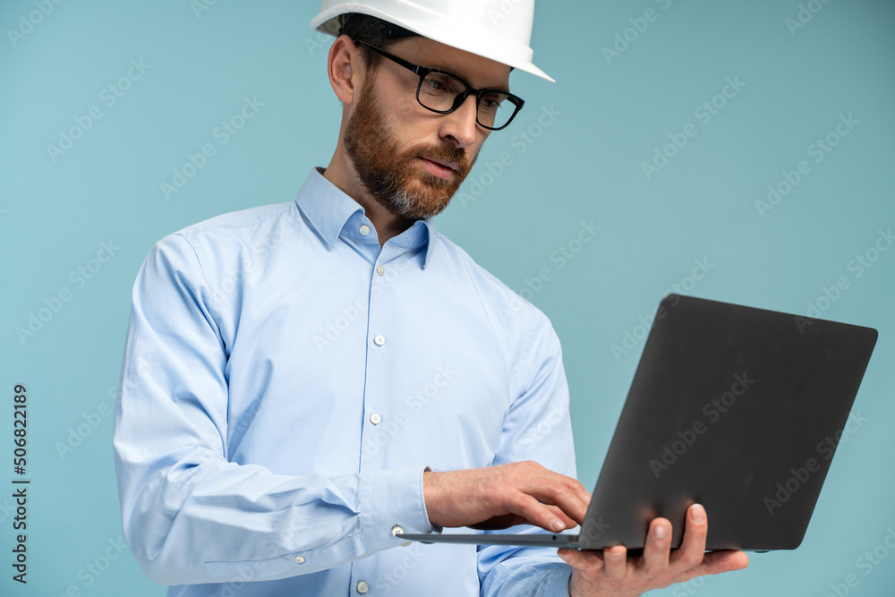 Young architect man with concentrated face in builder safety helmet looking at the laptop screen and typing at the keyboard over isolated background. Engineer concept Young architect man with concentrated face in builder safety helmet looking at the laptop screen and typing at the keyboard over isolated background. Engineer concept