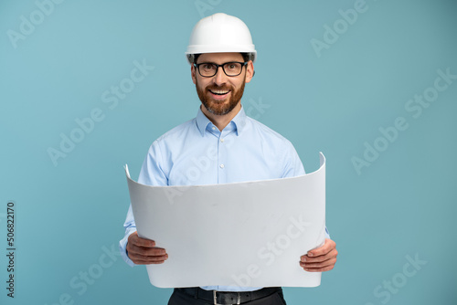 Architect man with cheerful face in builder safety helmet smiling while looking at the camera and holding blueprints over isolated background