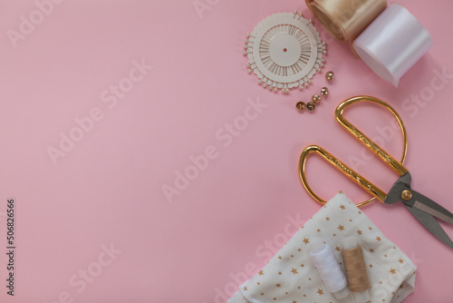The layout of sewing accessories and fabric on a pink background. Sewing threads, needles, fabric, buttons and scissors in gold and white. top view, flatlay with space for text