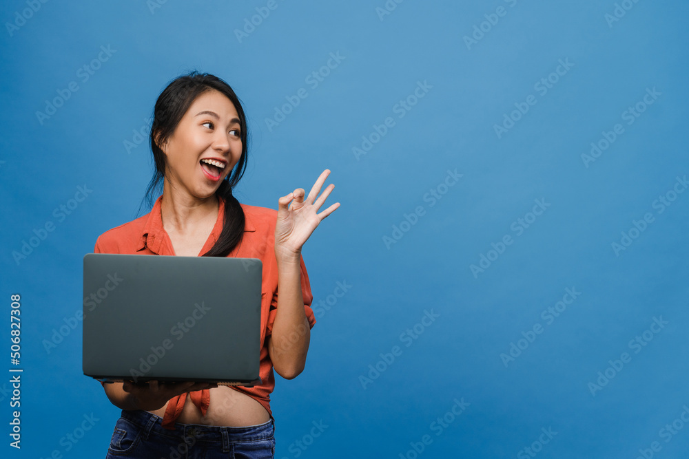 Young Asia lady using laptop with positive expression, smiles broadly, dressed in casual clothing feeling happiness and stand isolated on blue background. Happy adorable glad woman rejoices success. Young Asia lady using laptop with positive expression, smiles broadly, dressed in casual clothing feeling happiness and stand isolated on blue background. Happy adorable glad woman rejoices success.