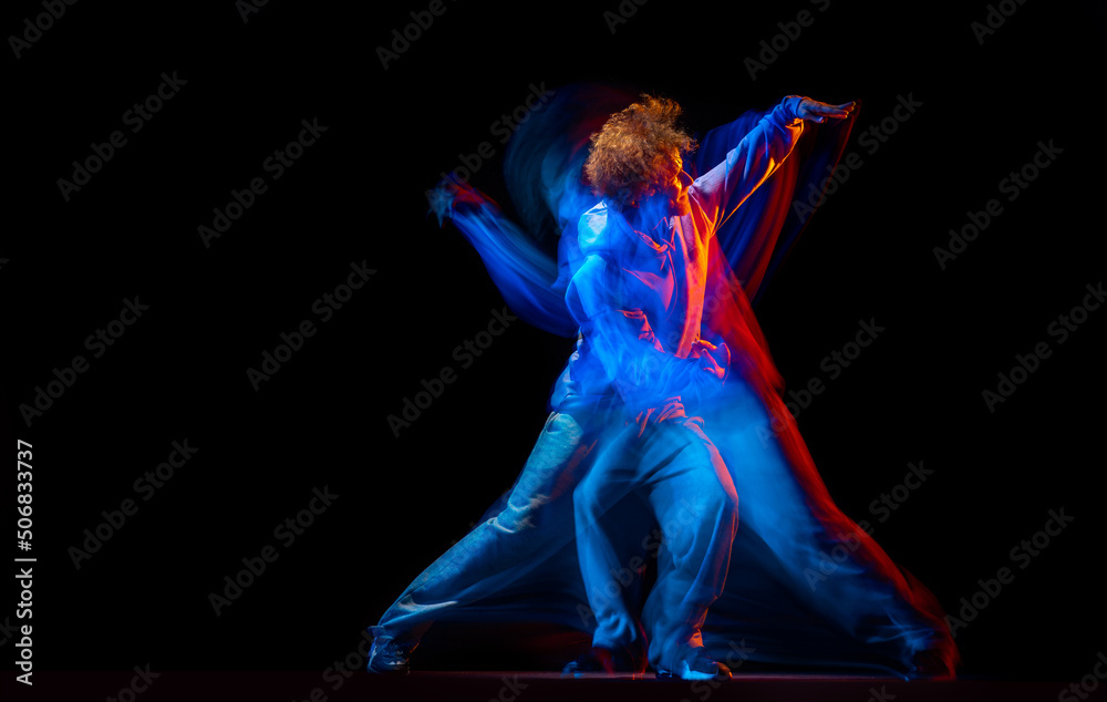 Stylish man dancing hip-hop in sportive style clothes isolated on dark background at dance hall in mixed neon light. Youth culture, hip-hop, movement