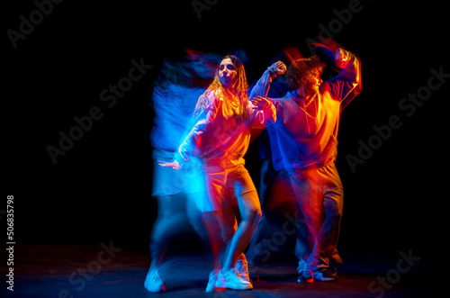 Young man and woman dancing hip-hop in sportive style clothes on dark background at dance hall in mixed neon light. Youth culture, hip-hop, movement