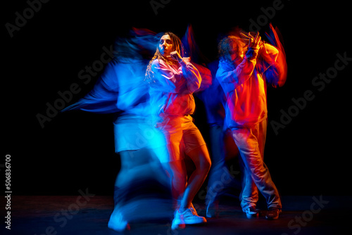 Dynamic portrait of stylish man and girl dancing hip-hop in sportive style clothes on dark background at dance hall in mixed neon light. Youth culture, breakdance, movement