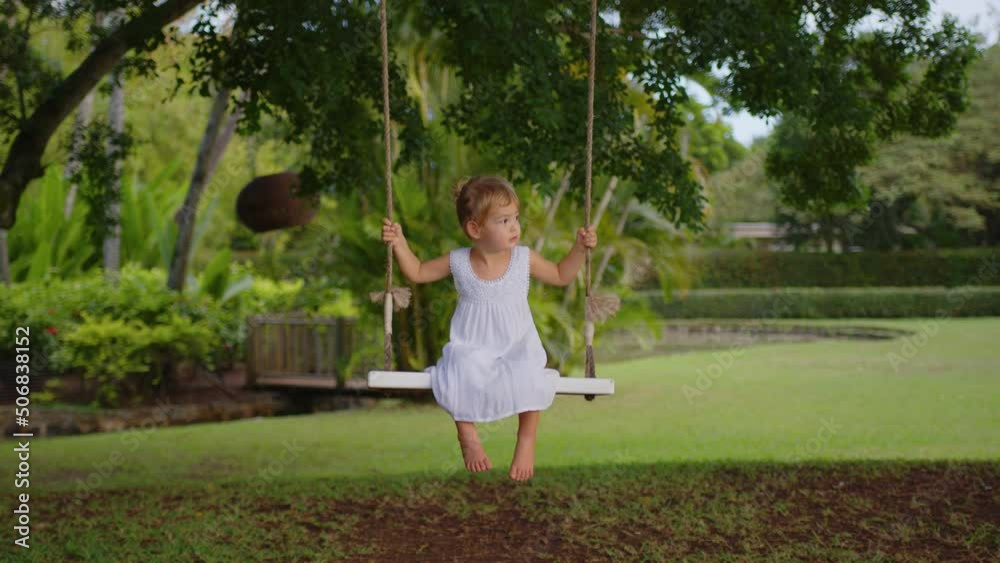 A little girl in a white dress rides on a rope swing in the park. The concept of outdoor recreation A little girl in a white dress rides on a rope swing in the park. The concept of outdoor recreation