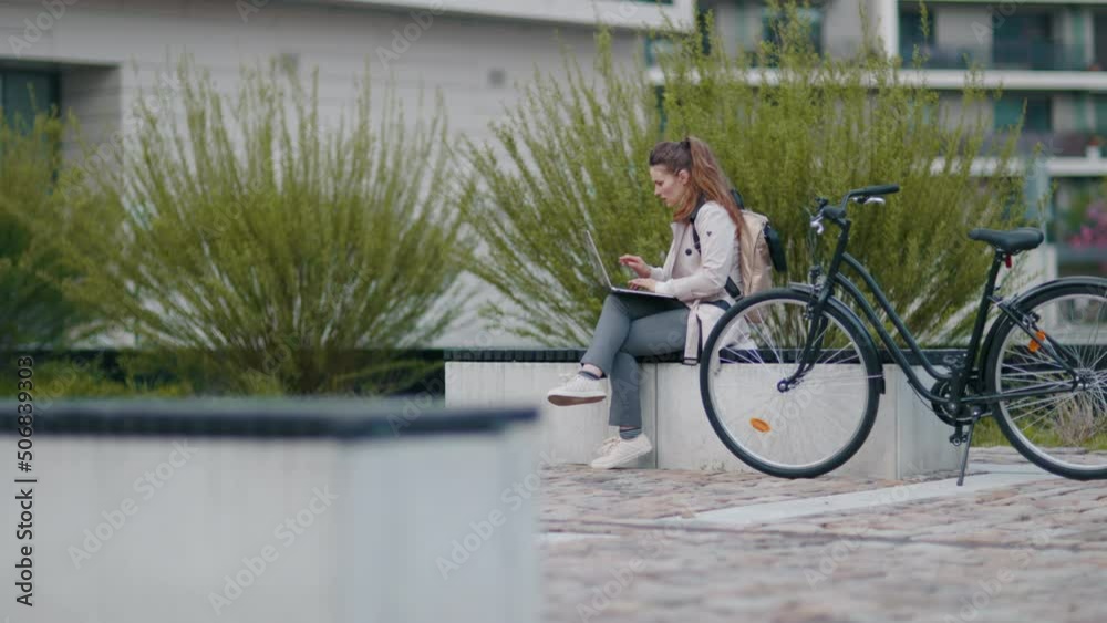 stylish woman in beige trench coat with bicycle and laptop sitting on the bench outside on the city street. stylish woman in beige trench coat with bicycle and laptop sitting on the bench outside on the city street.
