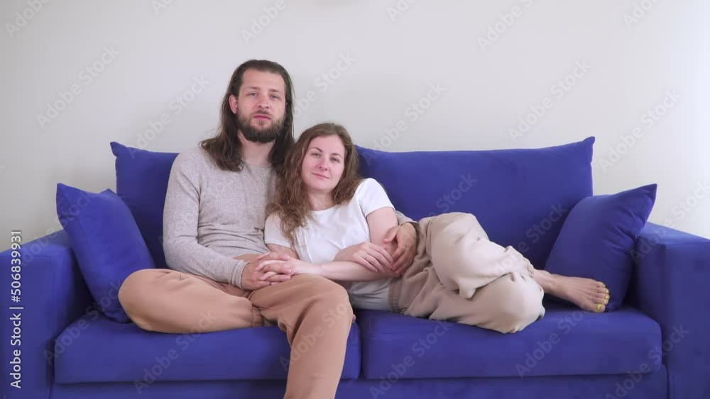 The couple is sitting on the couch hugging and kissing. Kiss of two lovers on a blue sofa. Caucasian man and woman in a cozy house spend time and weekends together. Be happy with each other. Smile. The couple is sitting on the couch hugging and kissing. Kiss of two lovers on a blue sofa. Caucasian man and woman in a cozy house spend time and weekends together. Be happy with each other. Smile.