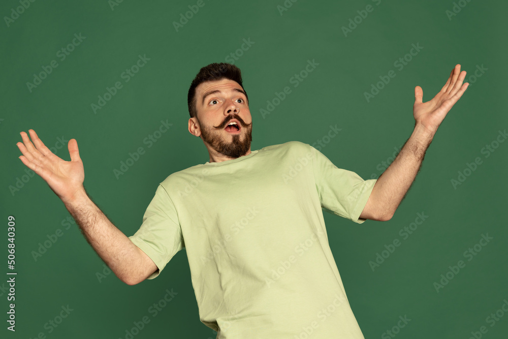 Portrait of young man with stylish moustache showing showing shocked expression, spreading hands isolated over green studio background