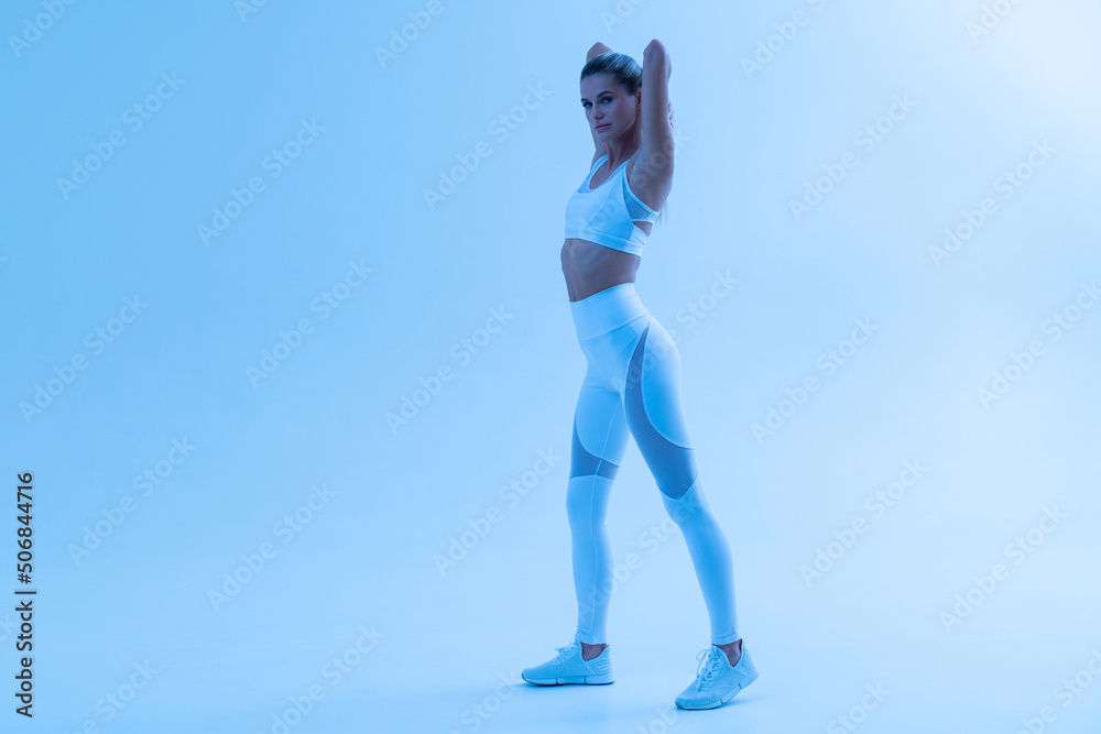 Beautiful slim female in sportswear standing in blue neon light with hands behind head