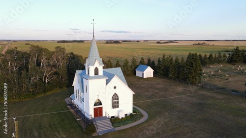 An ancient white church with a blue roof standing isolated in Canada's rural countryside. Wide angle aerial orbit photo